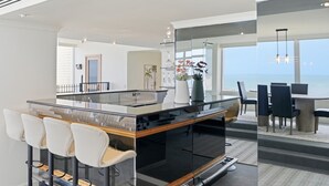 A modern kitchen with a central island, bar stools, and a view of the ocean. - The Penthouse | Spa, Sauna, Gym, 360° Views, 4BR (Glenelg North)