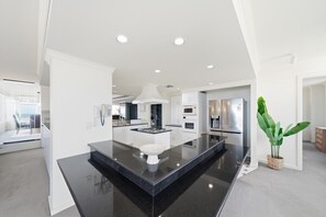 A modern kitchen with a central island, built-in appliances, and a large potted plant. - The Penthouse | Spa, Sauna, Gym, 360° Views, 4BR (Glenelg North)