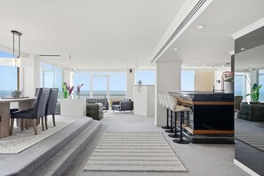 A modern interior with a dining area, bar, and seating by a large window overlooking the ocean. - The Penthouse | Spa, Sauna, Gym, 360° Views, 4BR (Glenelg North)
