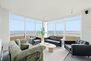 Ein geräumiges Wohnzimmer mit großen Fenstern, einem grünen Sofa, einem schwarzen Ledersessel und einem Couchtisch.