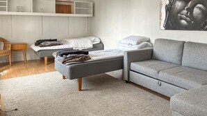 Quarto com sofá cinza, cama com colchão branco, mesa de cabeceira de madeira e prateleira na parede com objetos decorativos.