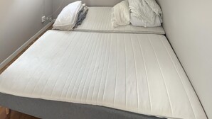 Uma cama com colchão branco e roupa de cama cinza, uma tomada na parede e um cabo correndo ao longo da parede.