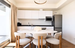A modern kitchen with white cabinets, a round dining table with chairs, and a large window with curtains. - Modern Apartment in Habitat Estepona (Estepona)
