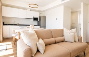 Uma sala moderna com um sofá bege, uma área de jantar com mesa branca e uma cozinha com armários e eletrodomésticos brancos.