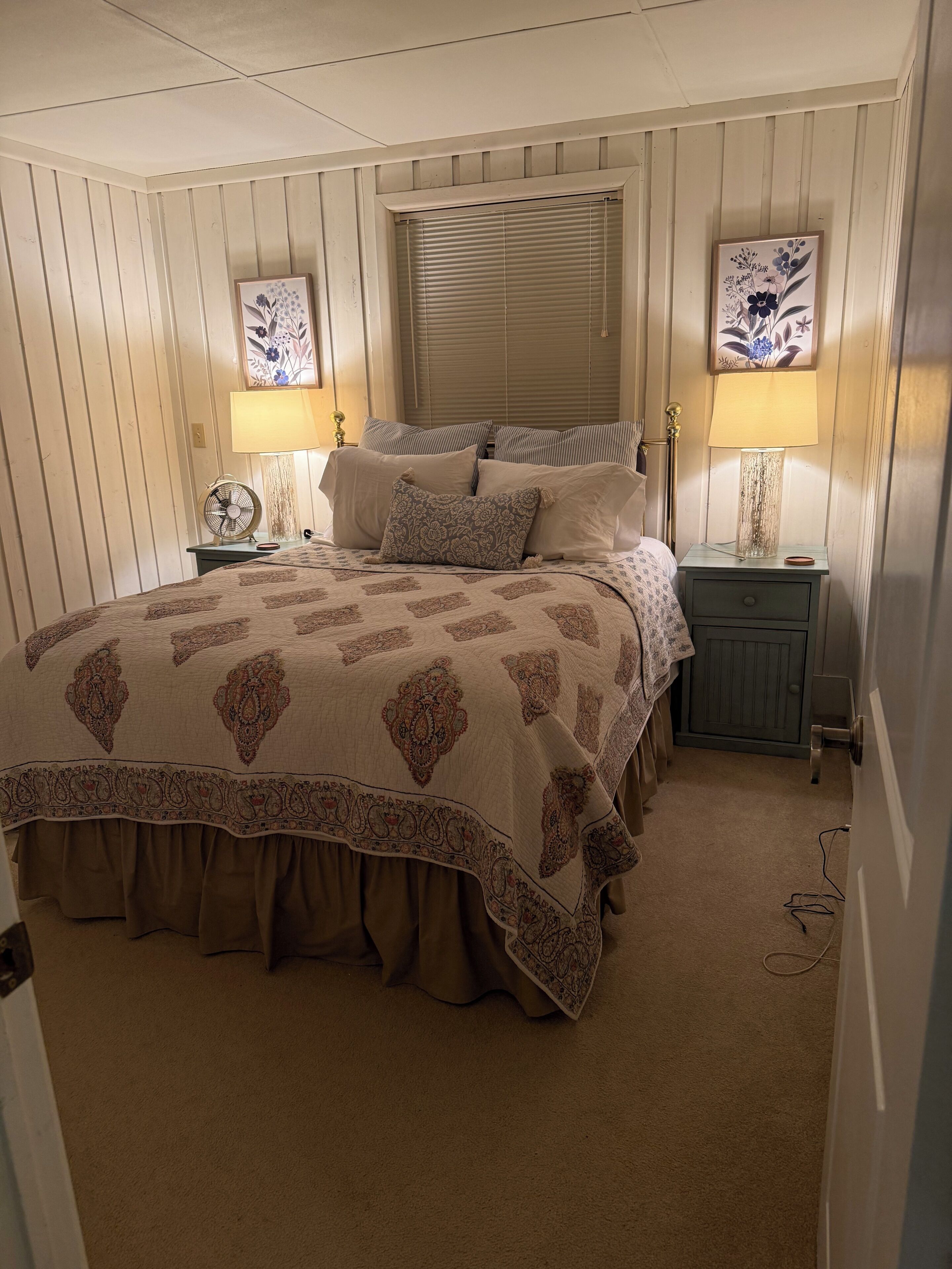 A bedroom with a large bed, two bedside lamps, a window with blinds, and a nightstand with a lamp.