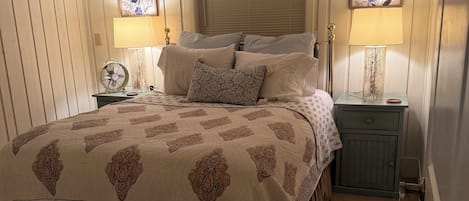 A bedroom with a large bed, two bedside lamps, a window with blinds, and a nightstand with a lamp.