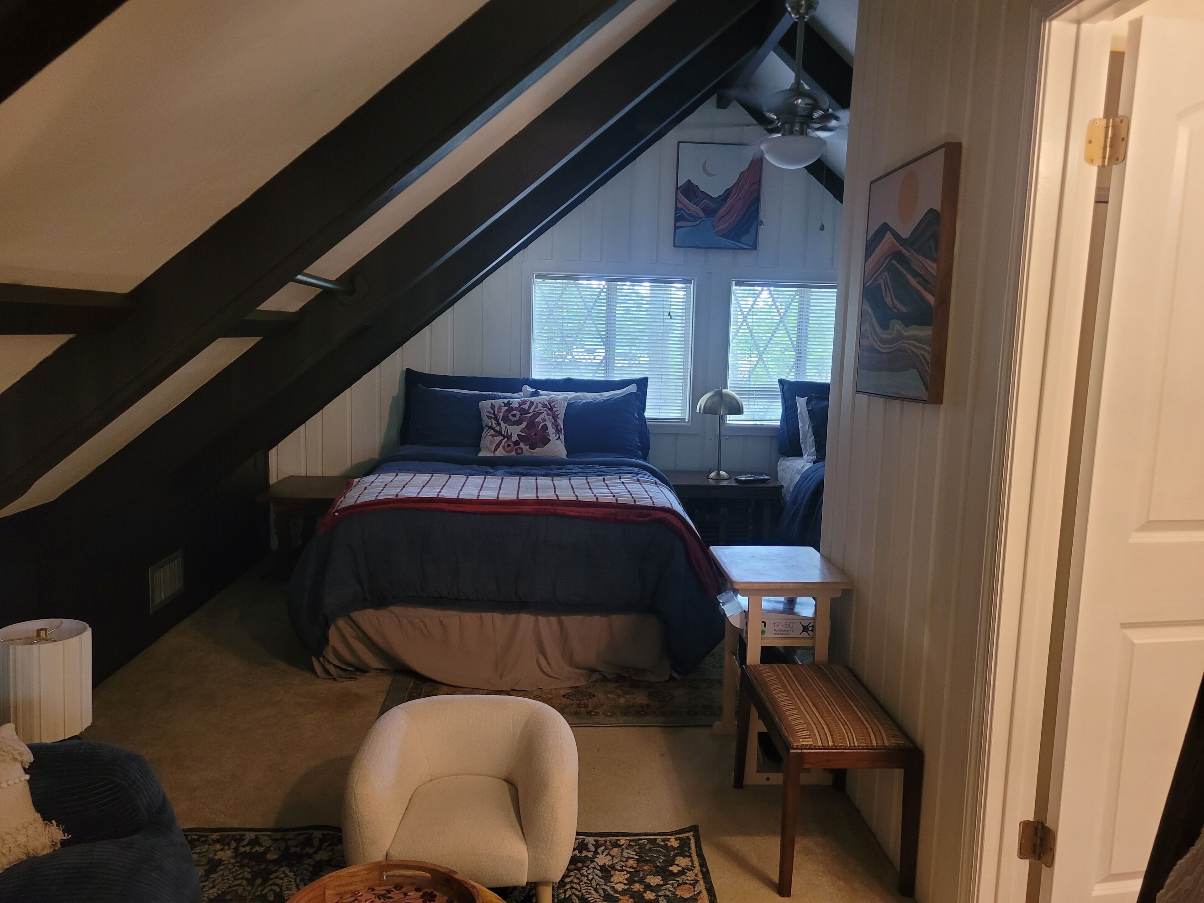A bedroom with a sloped ceiling, a bed with a red and white bedspread, a nightstand with a lamp, and a window with blinds.