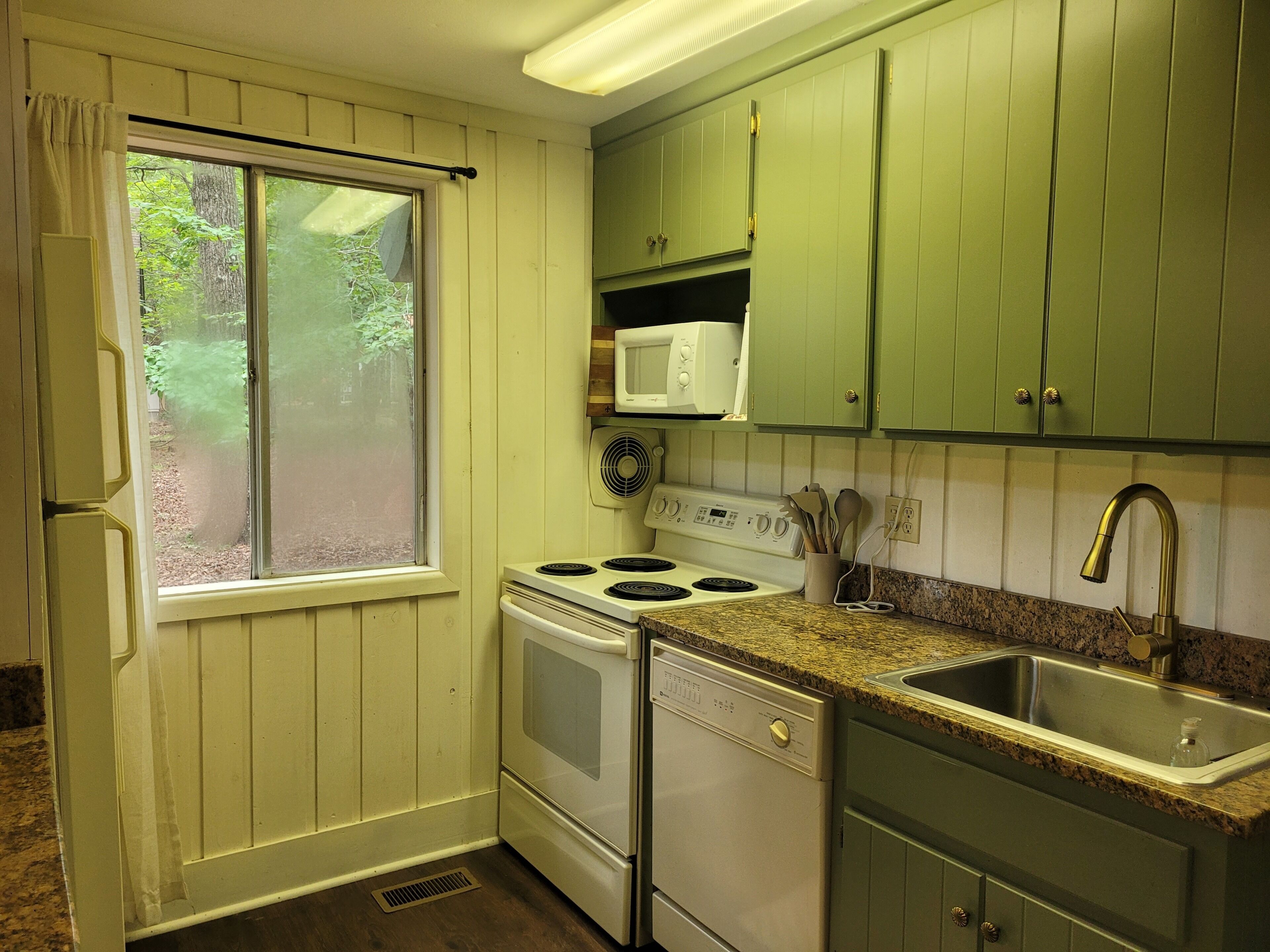 A compact kitchen with a window, white appliances, green cabinets, and a granite countertop.
