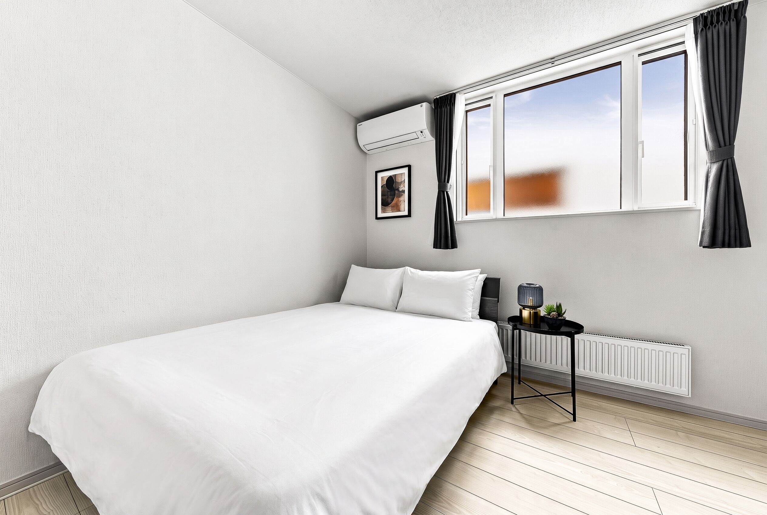 A bedroom with a bed, a radiator, a window with curtains, and a small table with a lamp and a plant.