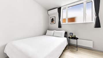 A bedroom with a bed, a radiator, a window with curtains, and a small table with a lamp and a plant.