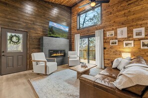 A cozy cabin interior with a fireplace, wooden walls, a large window, and a comfortable seating area. - Autumn Escape at Wears Valley • Hot Tub and Fireplace (Sevierville)