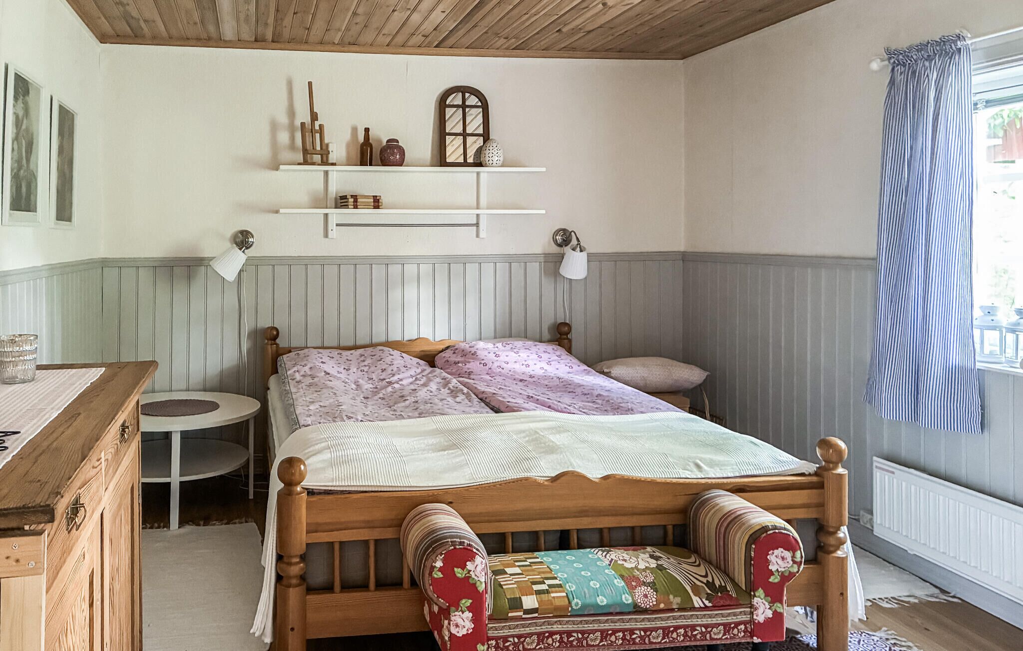 Une chambre à coucher avec un lit en bois, une table de chevet, une petite table ronde, une étagère avec des objets décoratifs et une fenêtre avec des rideaux.