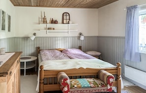 Un dormitori amb un llit de fusta, una taula de nit, una taula rodona petita, una estanteria amb objectes decoratius i una finestra amb cortines.