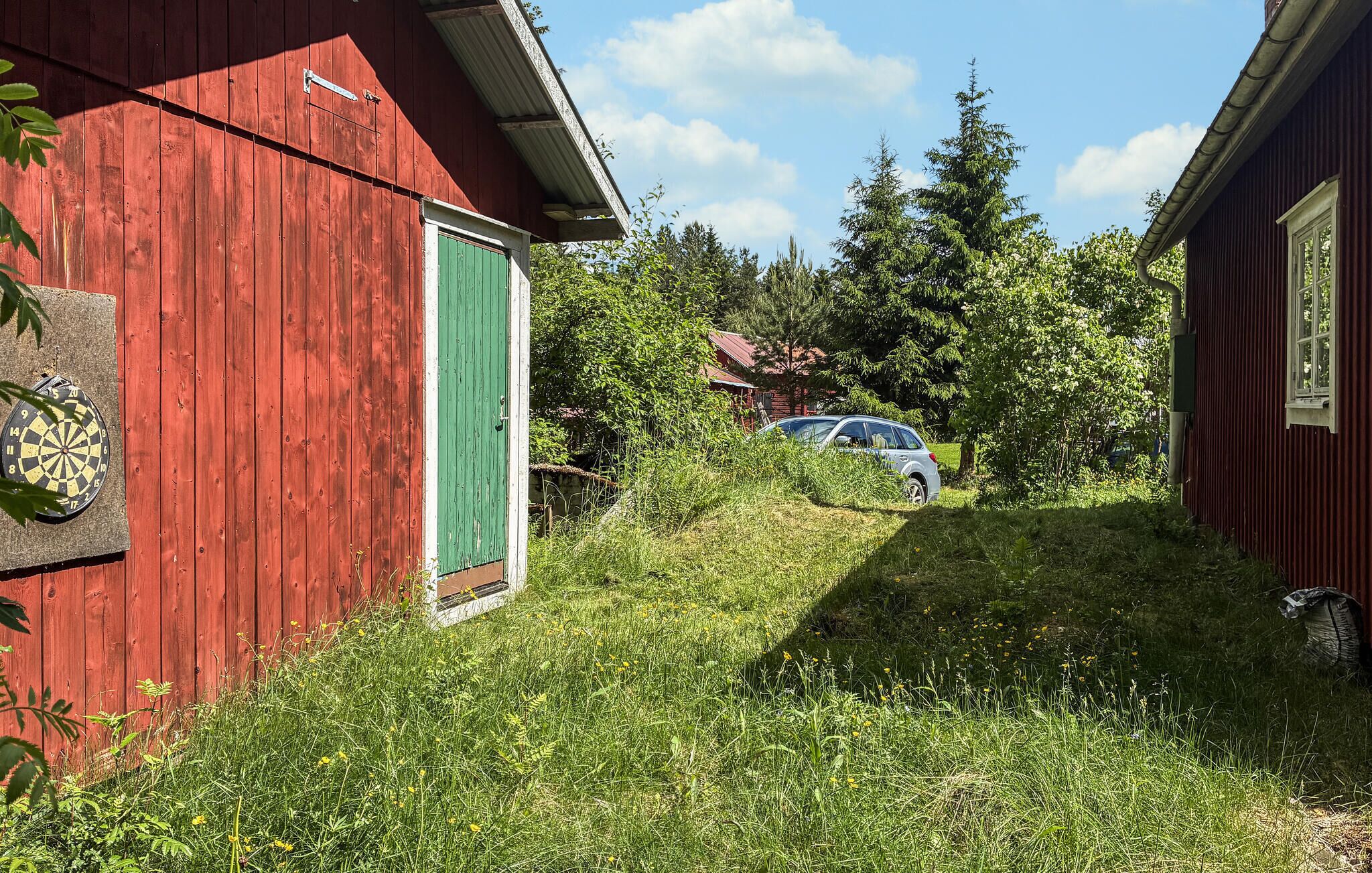 Un bâtiment en bois rouge avec une porte verte, une voiture garée à l’arrière-plan et une zone herbeuse avec des arbres.