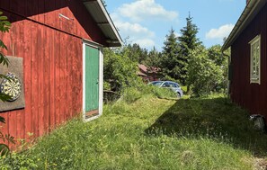 A red wooden building with a green door, a car parked in the background, and a grassy area with trees.