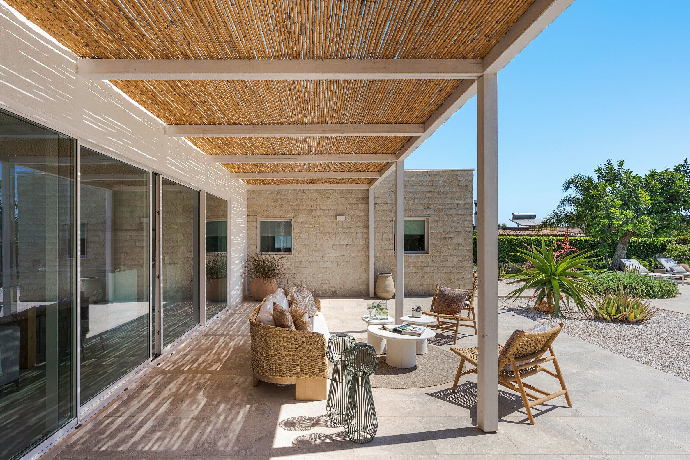 A covered outdoor seating area with wicker furniture, a glass wall, and a stone wall.