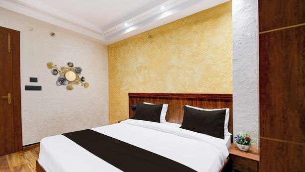A hotel room with a large bed, wooden headboard, a decorative wall piece, and a wooden nightstand with a plant. - Super Hotel O Prayagraj Junction (Prayagraj)