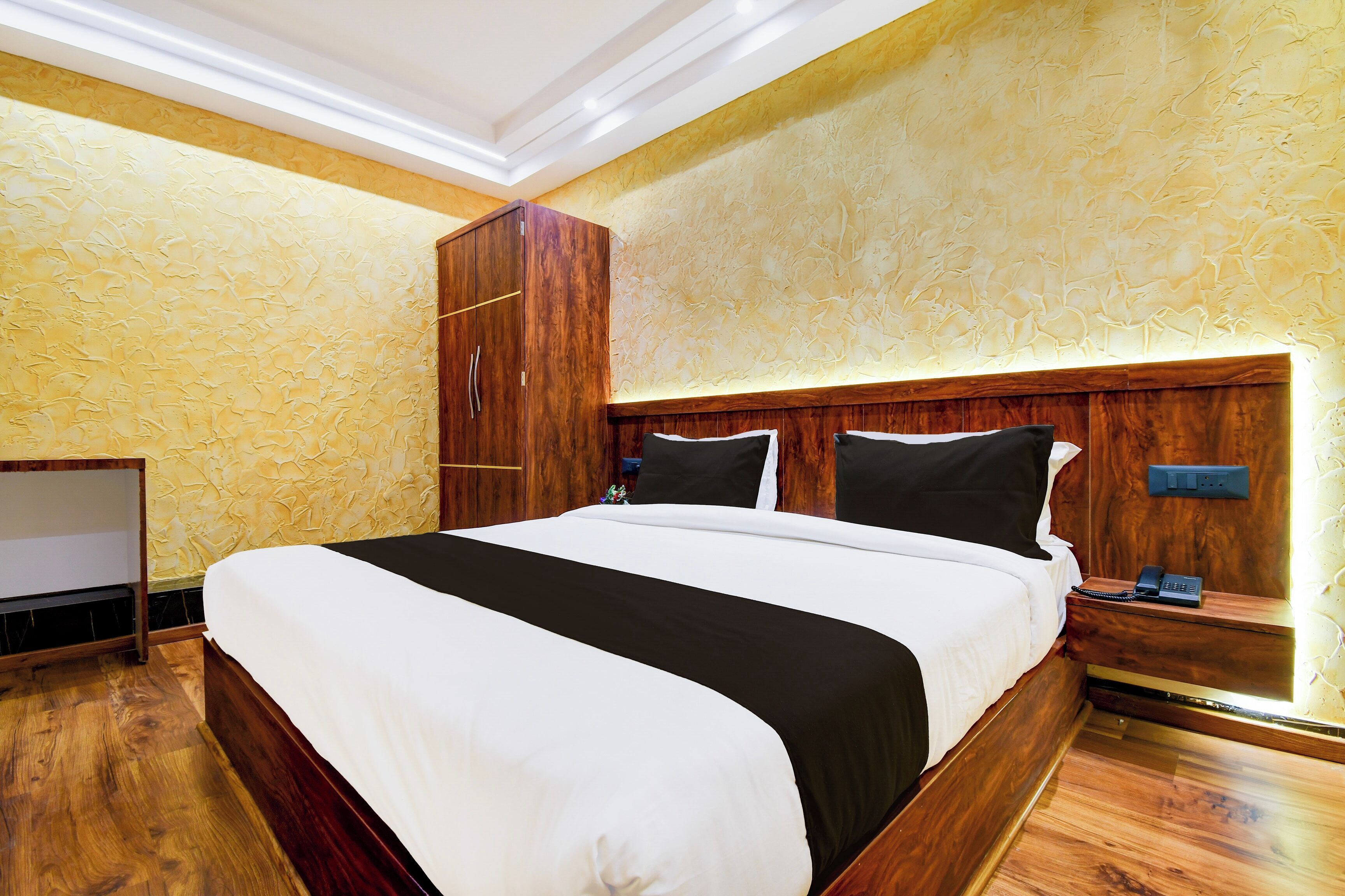 Une chambre d’hôtel avec un grand lit, une tête de lit en bois, une armoire, une table de chevet avec un téléphone et un luminaire fixé au mur.