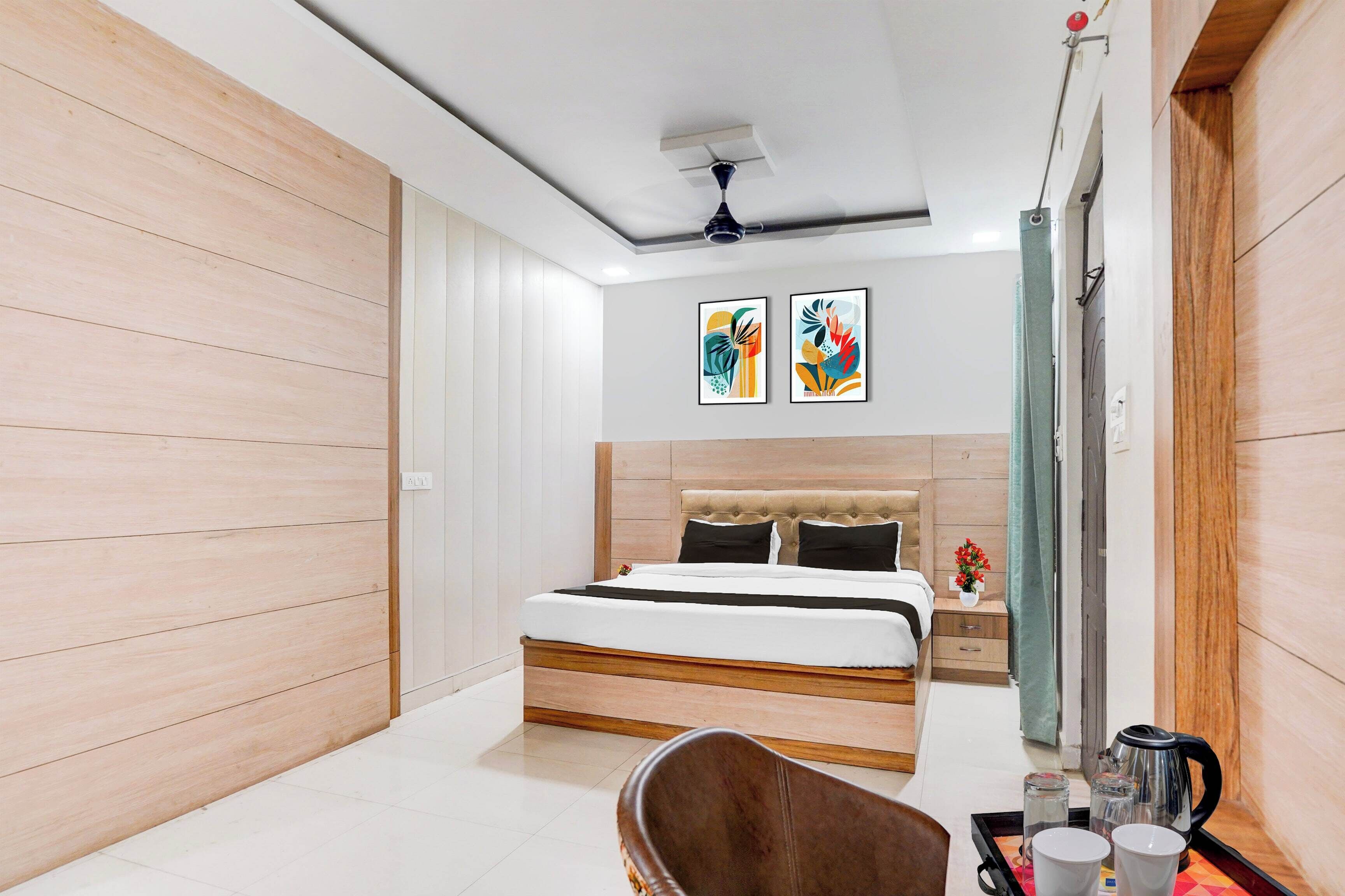 Une chambre moderne avec une tête de lit en bois, un lit recouvert de linge de lit blanc, deux œuvres d’art abstraites encadrées, un ventilateur de plafond et une petite table sur laquelle se trouvent une bouilloire et des tasses.