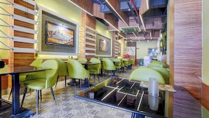 Interior de restaurante com cadeiras estofadas verdes, soalho estampado e um quadro grande emoldurado na parede.