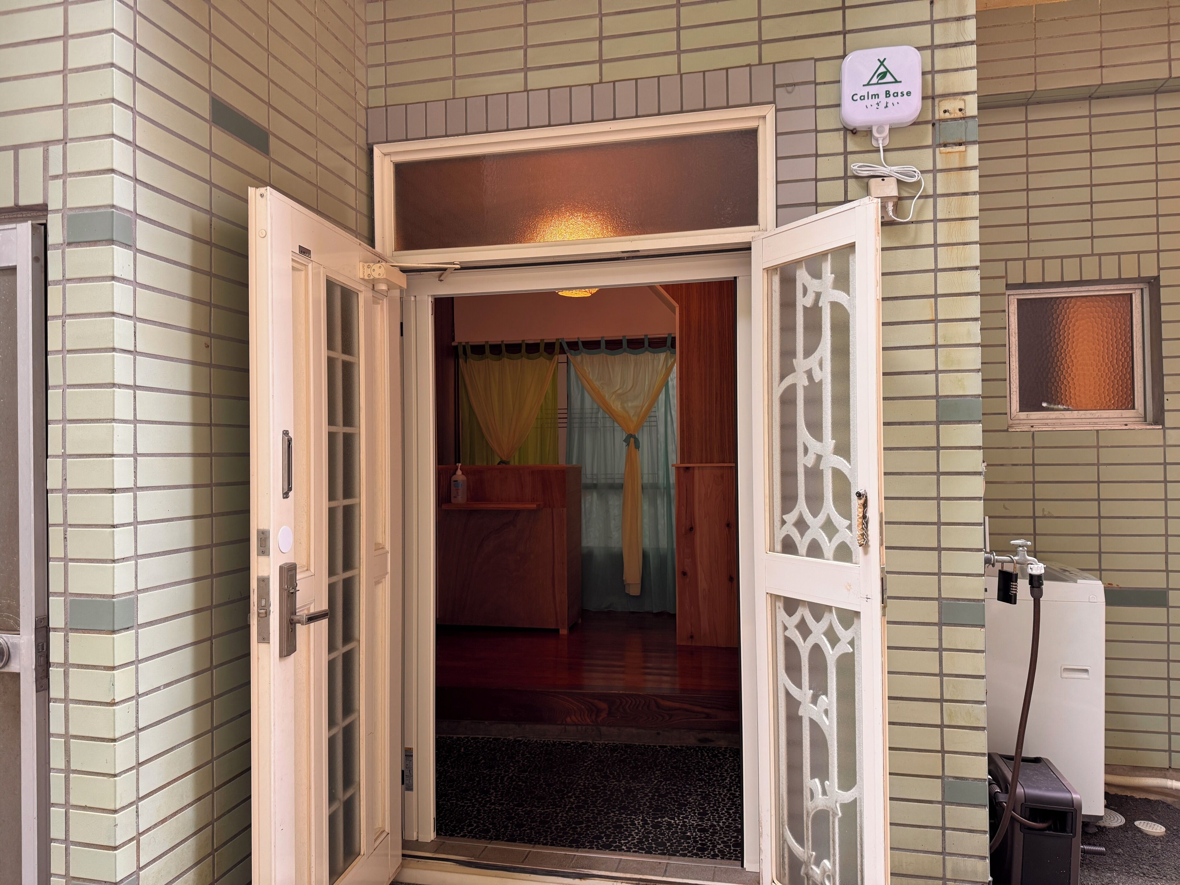 A hotel entrance with a tiled wall, a door with a decorative glass panel, a small indoor garden, and a water dispenser.