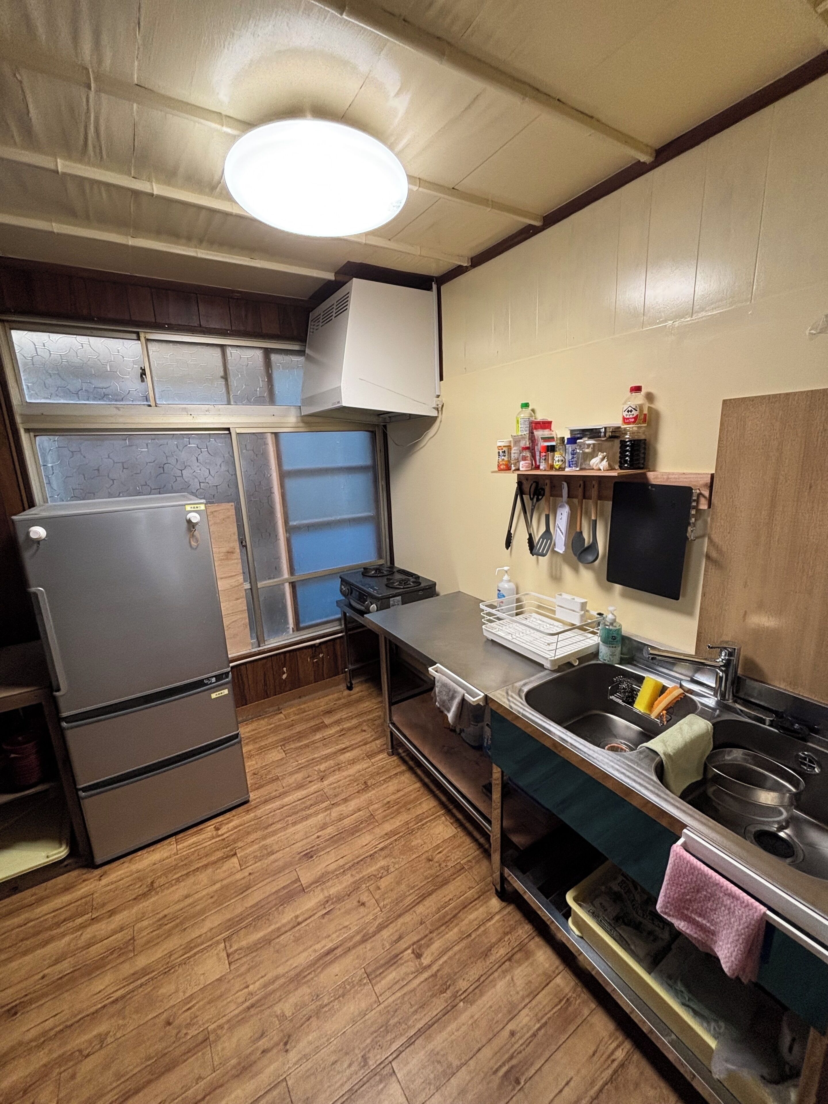 A compact kitchen with a refrigerator, stove, and sink. There is a window with frosted glass, a shelf with kitchen items, and a wall-mounted TV.