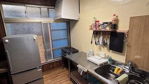 A compact kitchen with a refrigerator, stove, and sink. There is a window with frosted glass, a shelf with kitchen items, and a wall-mounted TV. - Calmbase Nishiizu Izayoi (Numazu)