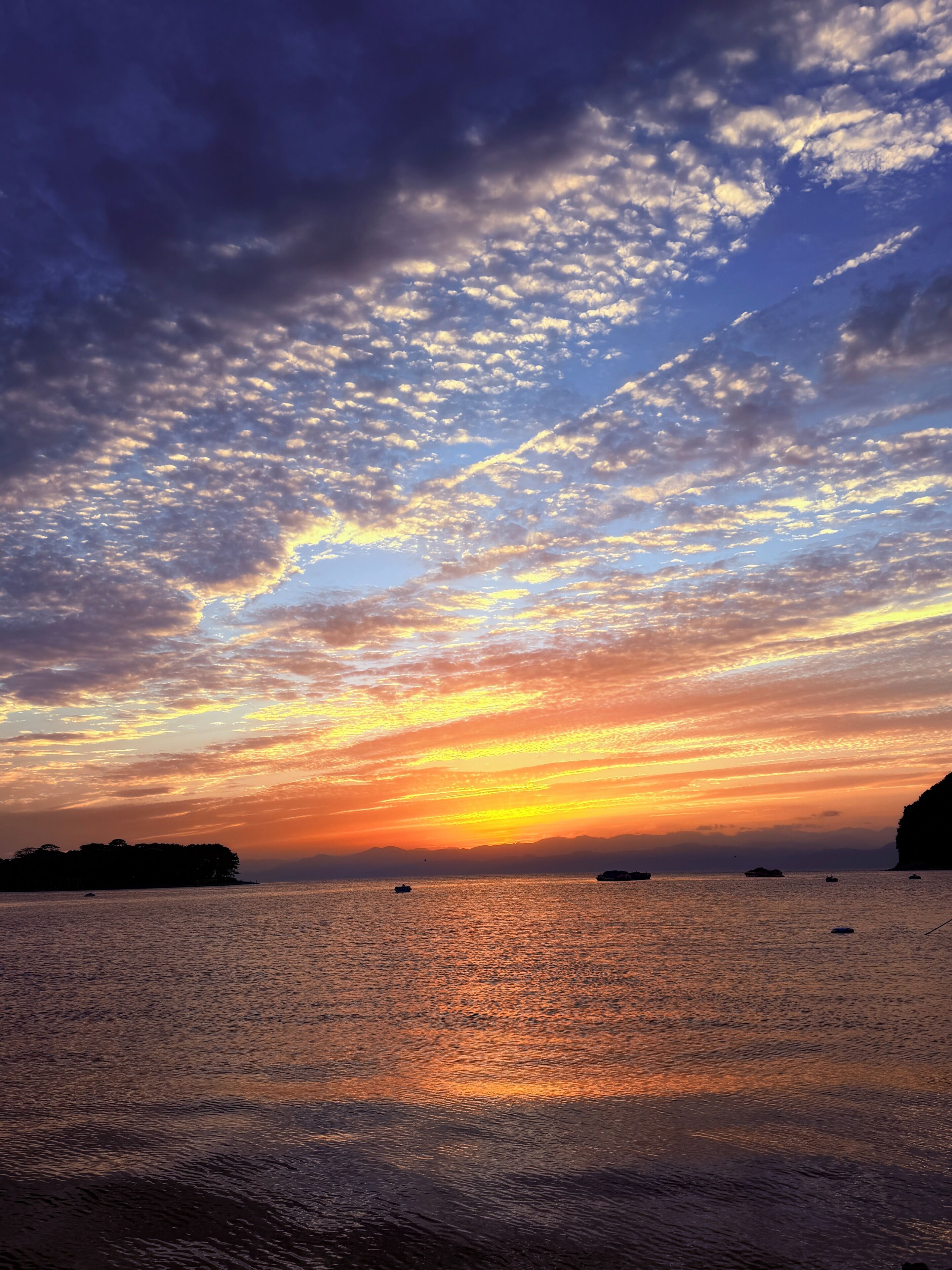A sunset over a calm sea with boats and distant land.