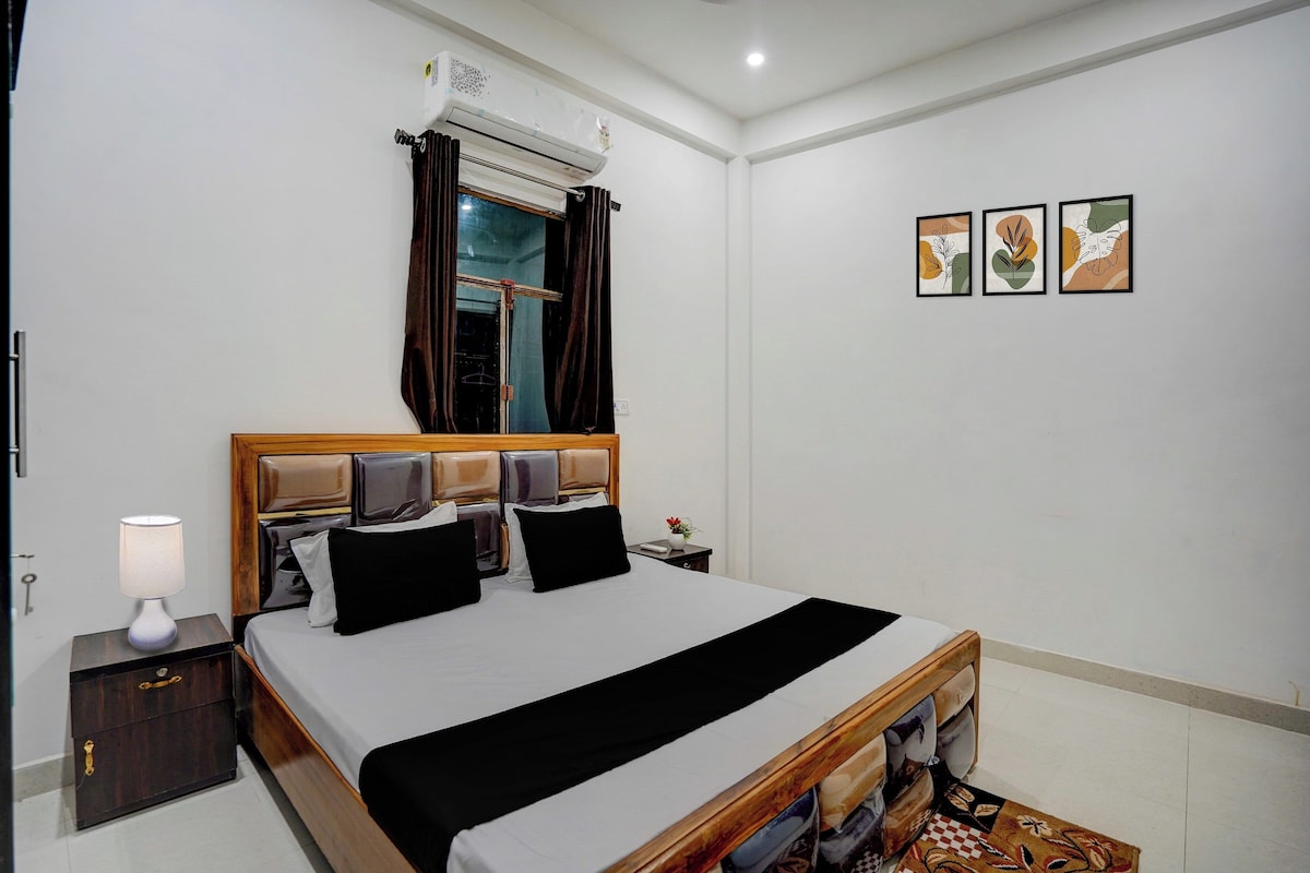Habitación de hotel con cama, mesita de noche, lámpara, aire acondicionado, ventana con cortinas y tres cuadros enmarcados en la pared.