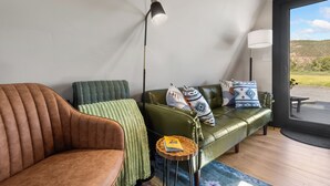 Ein modernes Wohnzimmer mit einem braunen Ledersofa, einem grünen Sofa mit gemusterten Kissen, einer Stehlampe und Blick durch eine Glastür.