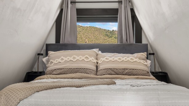 A bed with patterned bedding and a view of a hilly landscape through a window. - Whispering Pines A-Frame Retreat | Sleeps 2 | +AC (Ridgway)