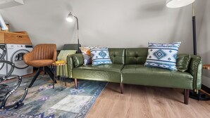 A modern living room with a green leather sofa, a patterned rug, a standing lamp, and a washing machine. - Whispering Pines A-Frame Retreat | Sleeps 2 | +AC (Ridgway)
