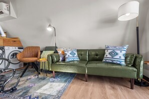 A modern living room with a green leather sofa, a patterned rug, a standing lamp, and a washing machine. - Whispering Pines A-Frame Retreat | Sleeps 2 | +AC (Ridgway)