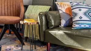 A green sofa with a patterned pillow, a brown chair, and a small wooden table with a yellow item on it. - Whispering Pines A-Frame Retreat | Sleeps 2 | +AC (Ridgway)