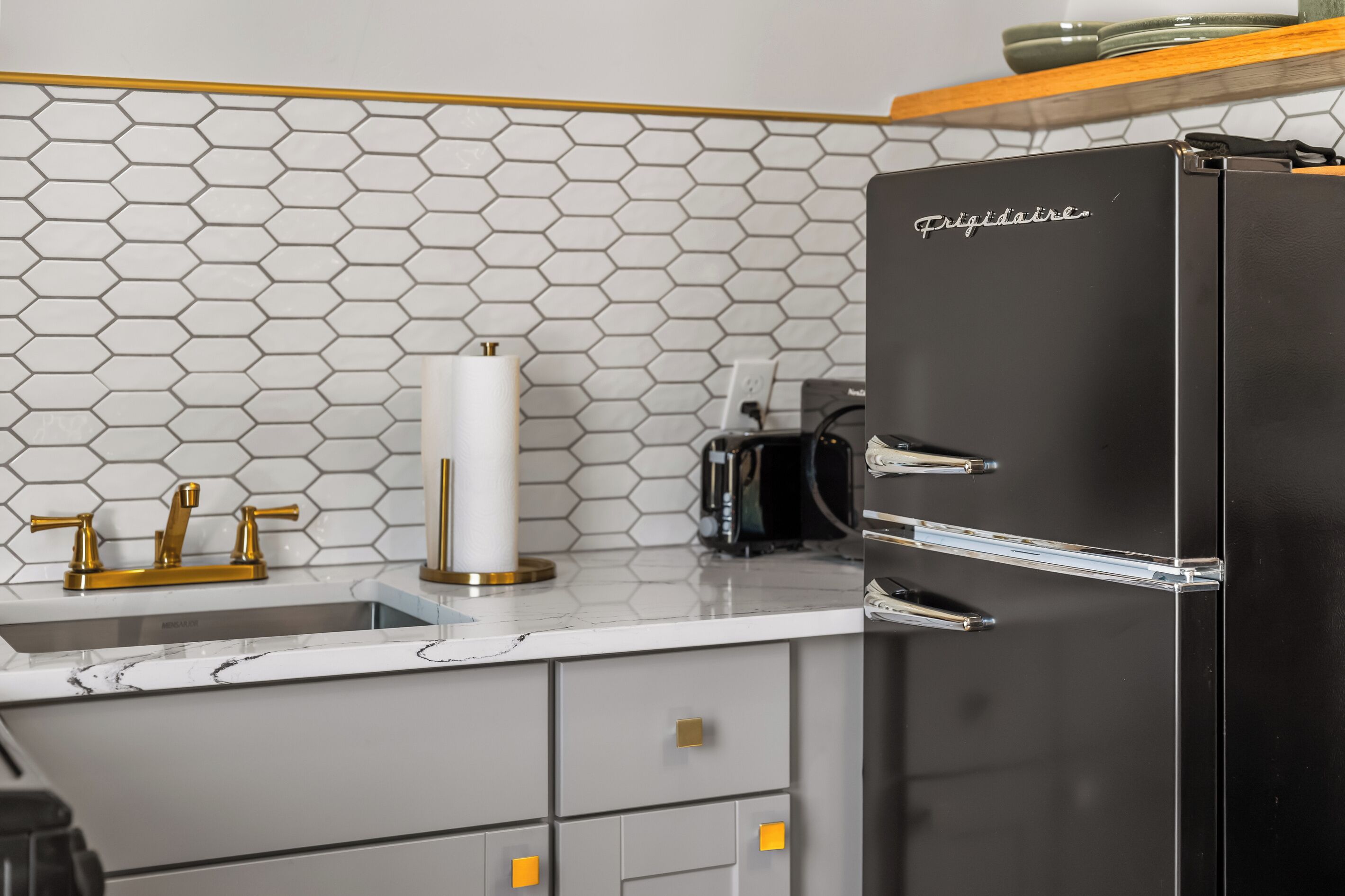 A kitchen with a black refrigerator, a sink with gold faucets, and a hexagonal tile backsplash.