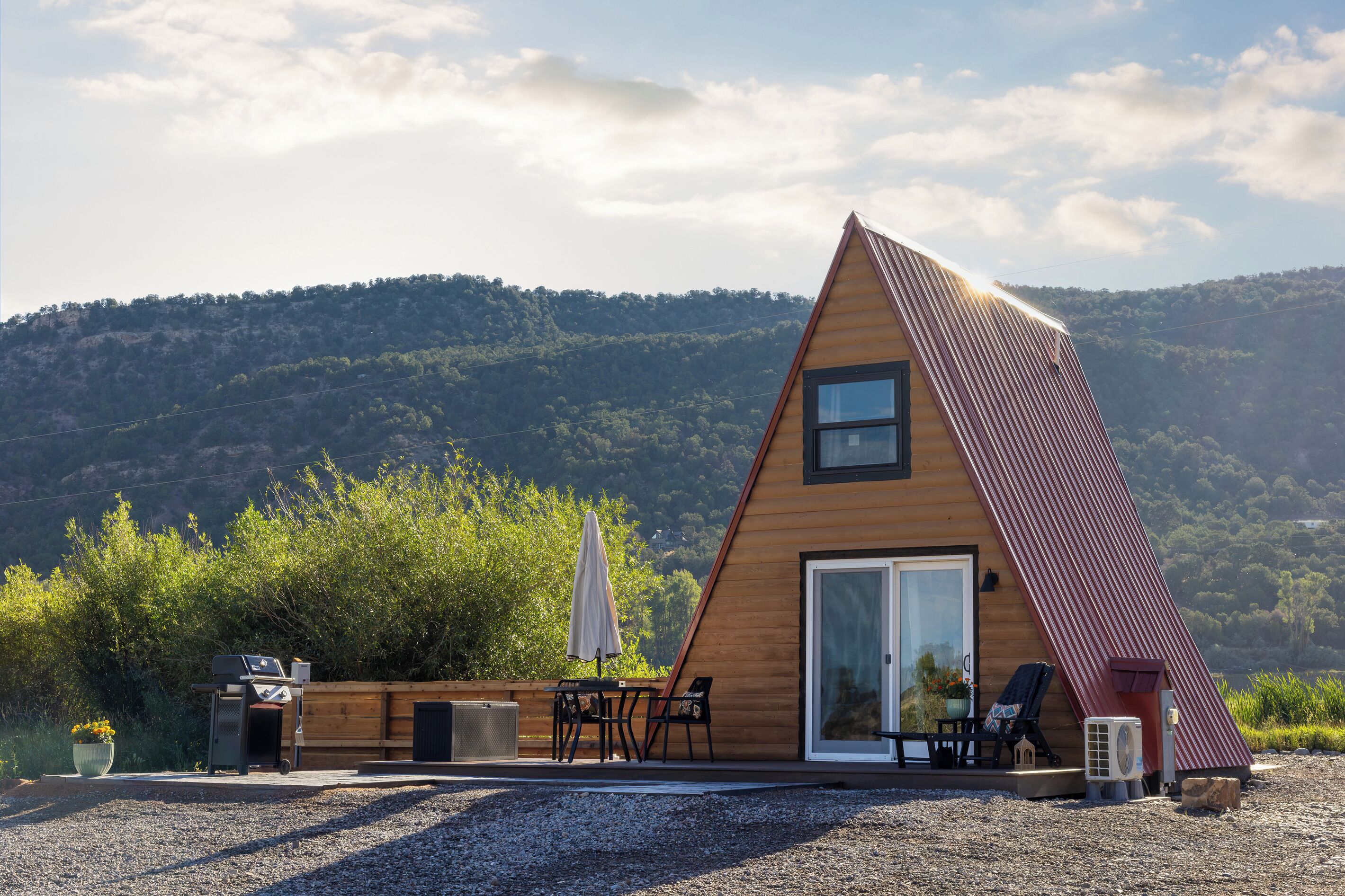 A modern, triangular wooden house with a large sliding glass door, outdoor seating, and a gravel driveway.