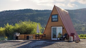 A modern, triangular wooden house with a large sliding glass door, outdoor seating, and a gravel driveway. - Whispering Pines A-Frame Retreat | Sleeps 2 | +AC (Ridgway)