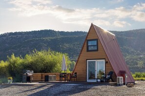 A modern, triangular wooden house with a large sliding glass door, outdoor seating, and a gravel driveway. - Whispering Pines A-Frame Retreat | Sleeps 2 | +AC (Ridgway)