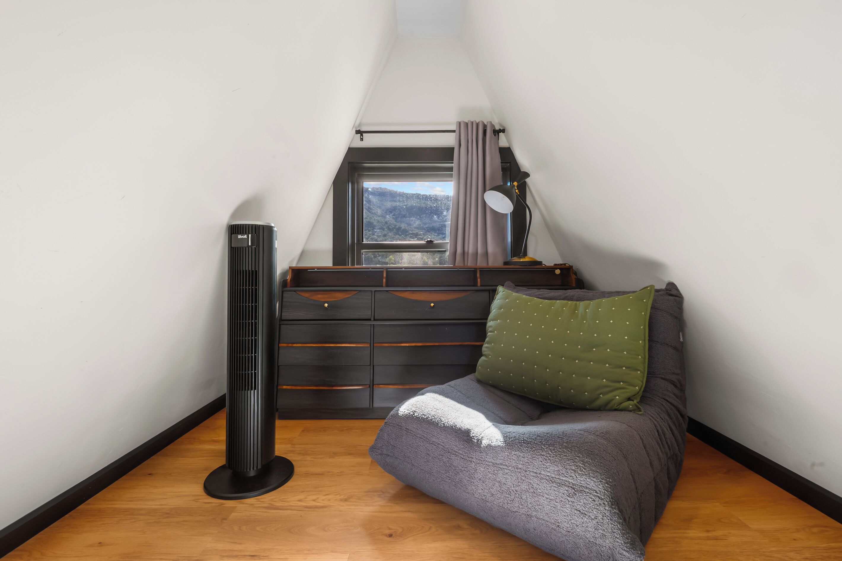 A small, cozy room with a sloped ceiling, a radiator, a bed with a green pillow, a desk with a lamp, and a view of a mountainous landscape through a window.
