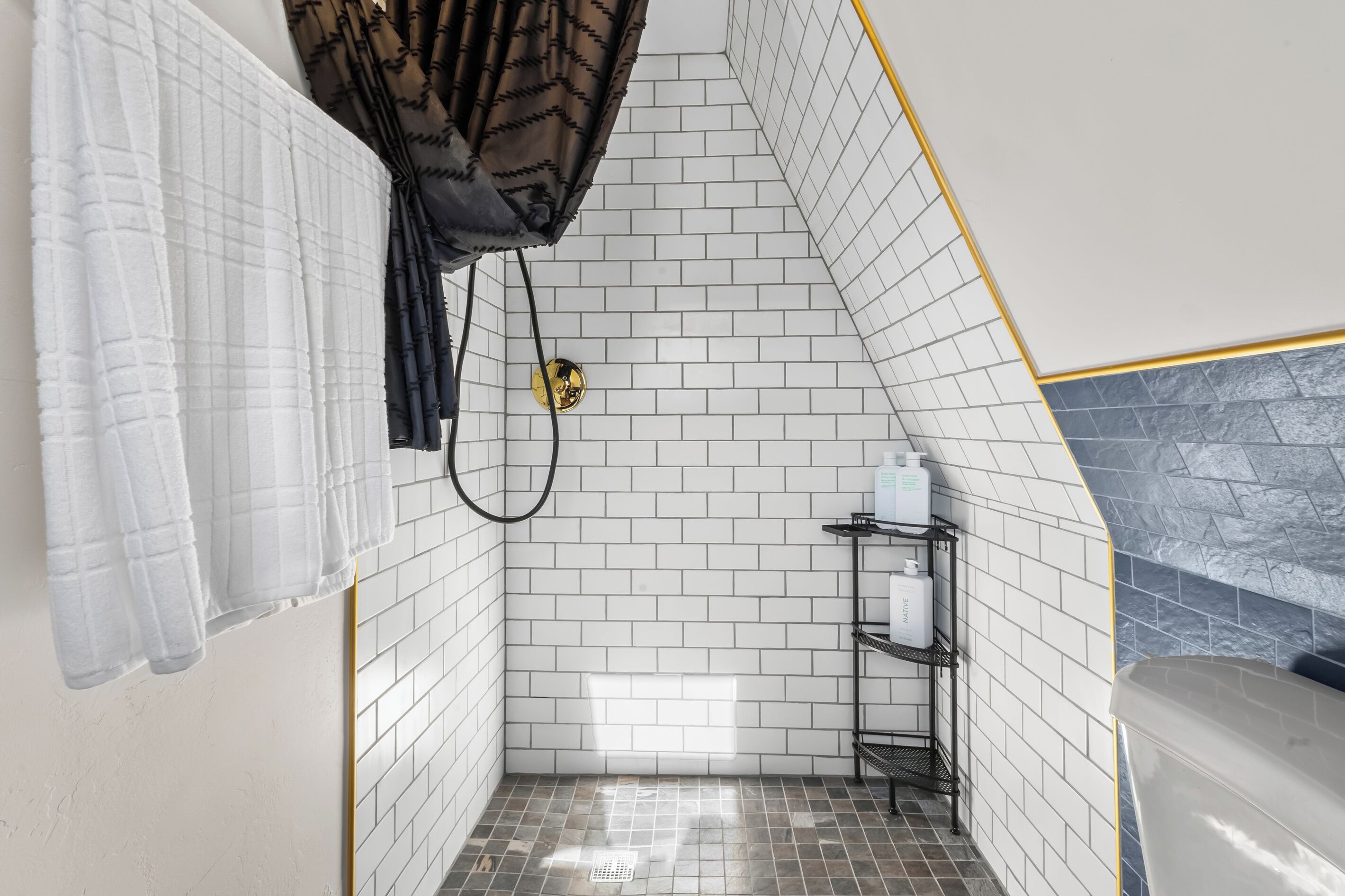 A bathroom with white tiled walls, a shower area, and a black metal shelf with toiletries.