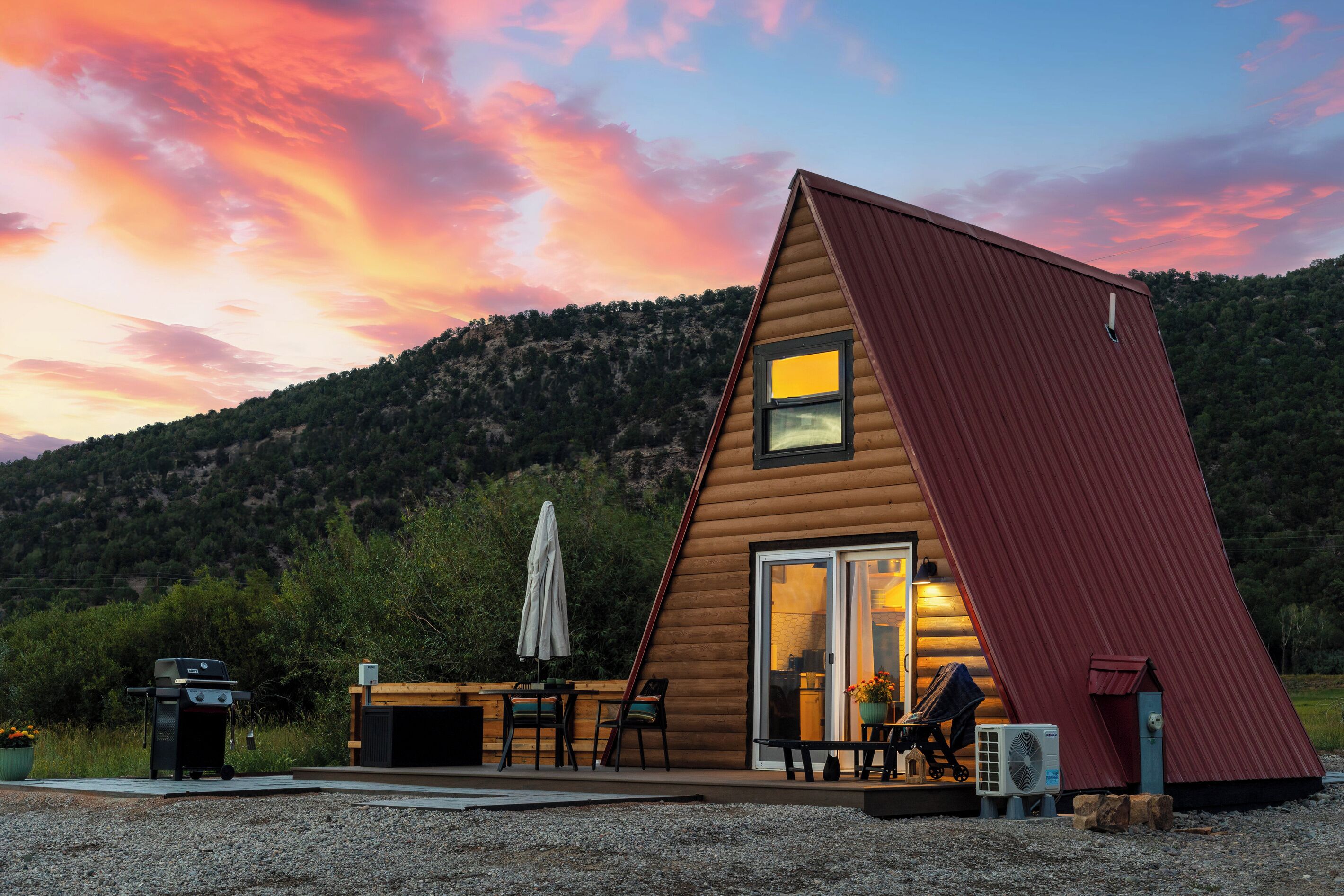 A wooden cabin with a sloped roof, a patio with a table and chairs, and a view of a forested hillside.