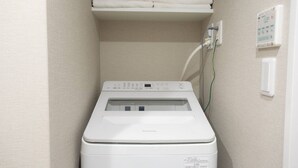 Een compacte, witte wasmachine in een kleine wasruimte, met een plank erboven vol opgevouwen handdoeken.