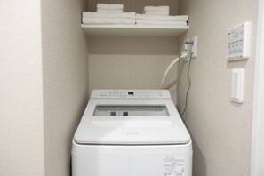 A compact, white washing machine in a small laundry room with a shelf above containing folded towels. - 1 min walk from sta./Excellent access to central (Osaka)