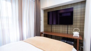 Una habitación de hotel con una cama, un televisor instalado en la pared, una mesita de noche con una tetera y una ventana con cortinas.