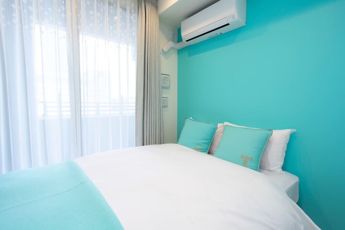 Ein Hotelzimmer mit einem Bett, einem Fenster mit durchscheinenden Vorhängen und einer Klimaanlage an der Wand.