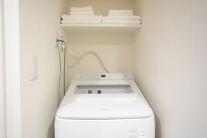 A compact, white front-loading washing machine in a small laundry room with a shelf above for folded towels. - 1 min walk from sta./Excellent access to central (Osaka)