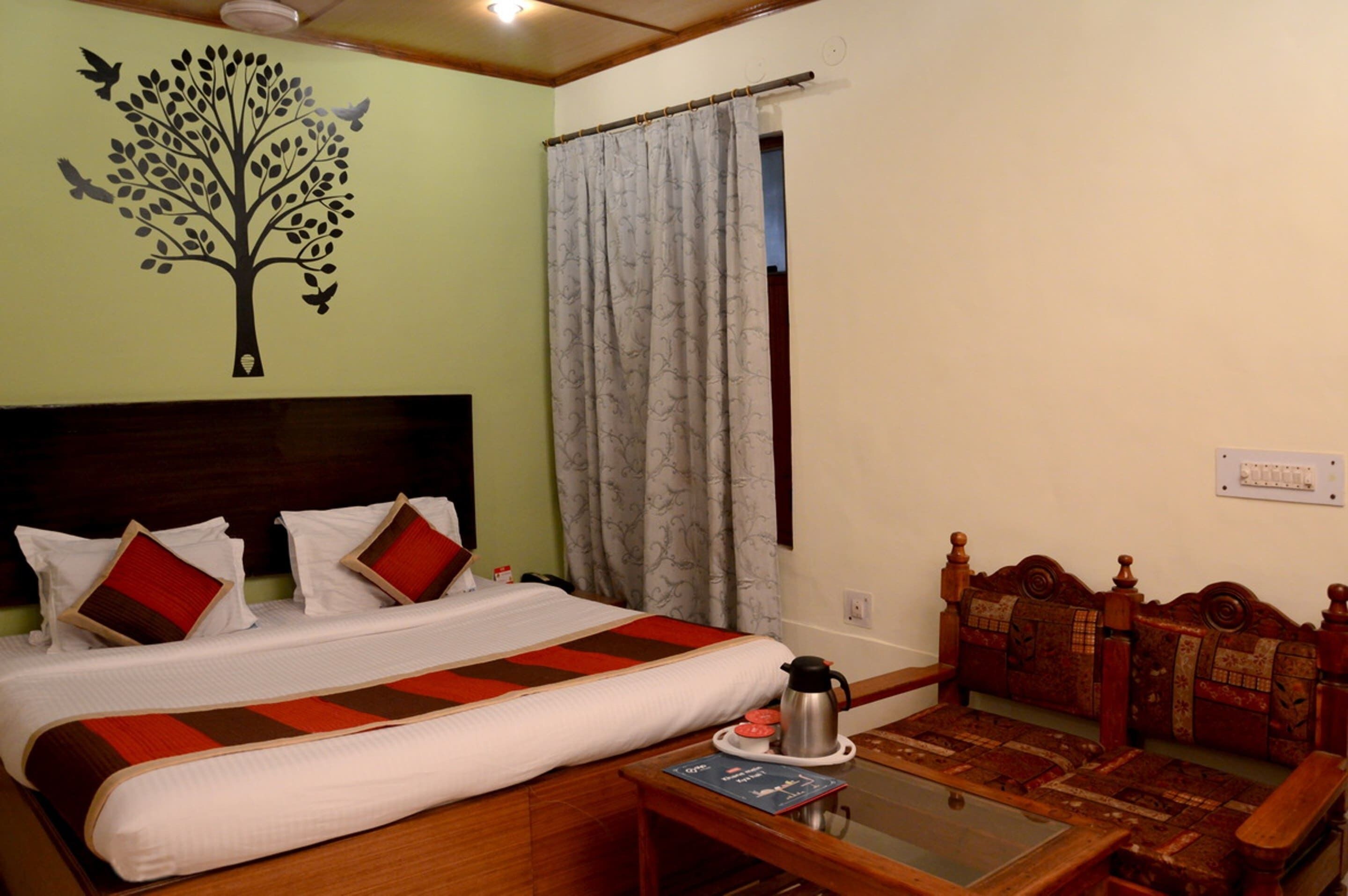 A hotel room with a bed, a wooden headboard, a bedside table with a book and a thermos, a window with curtains, and a wall decoration of a tree.