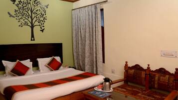 A hotel room with a bed, a wooden headboard, a bedside table with a book and a thermos, a window with curtains, and a wall decoration of a tree.