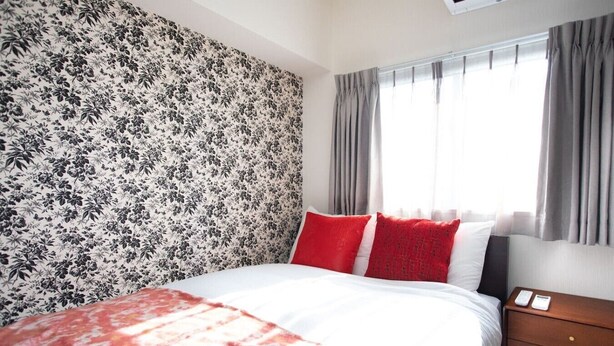 Una habitación de hotel con papel tapiz estampado con motivos florales, una cama con ropa de cama blanca y roja, y una mesita de noche de madera con un teléfono.