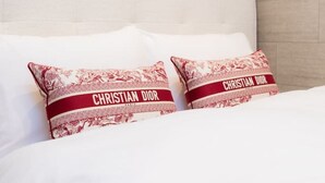 Ein Bett mit weißer Bettwäsche und zwei Kissen mit einem roten und weißen Muster sowie dem Text 'CHRISTIAN DIOR'.